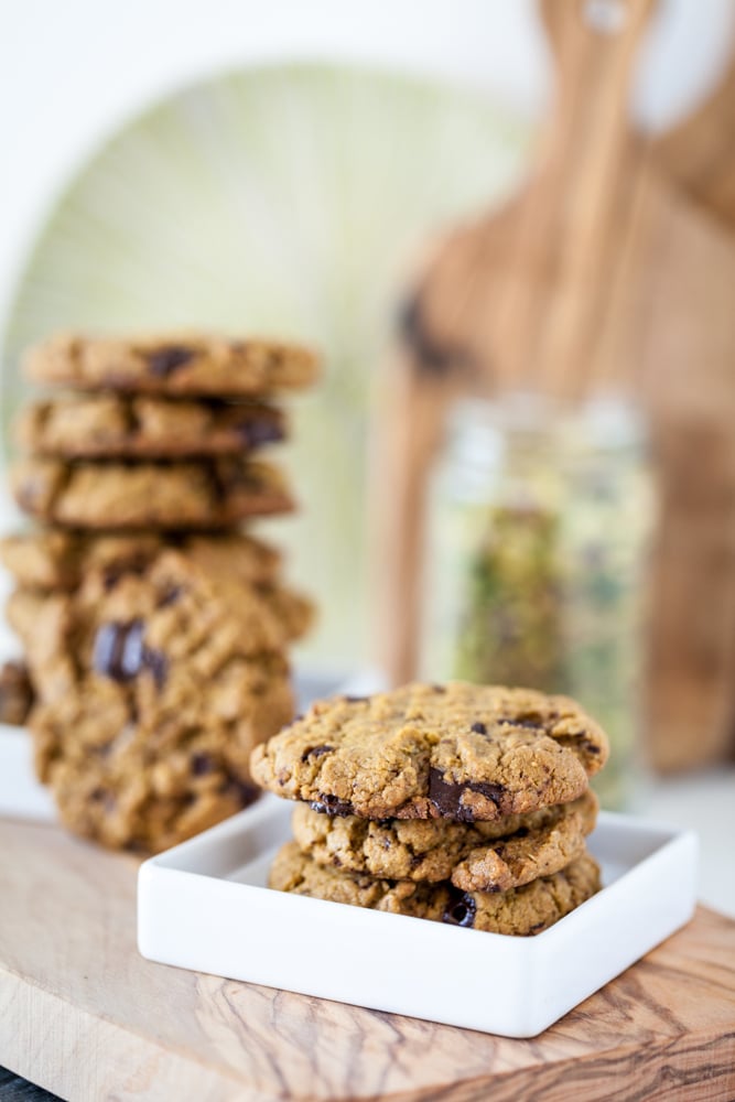 An easy Pistachio Cookies recipe with pistachio butter as the main ingredient and chocolate chunks for extra good flavor! Vegan, Gluten Free. pistachio cookies on white plate with stack of vegan cookies in bakground on wooden board