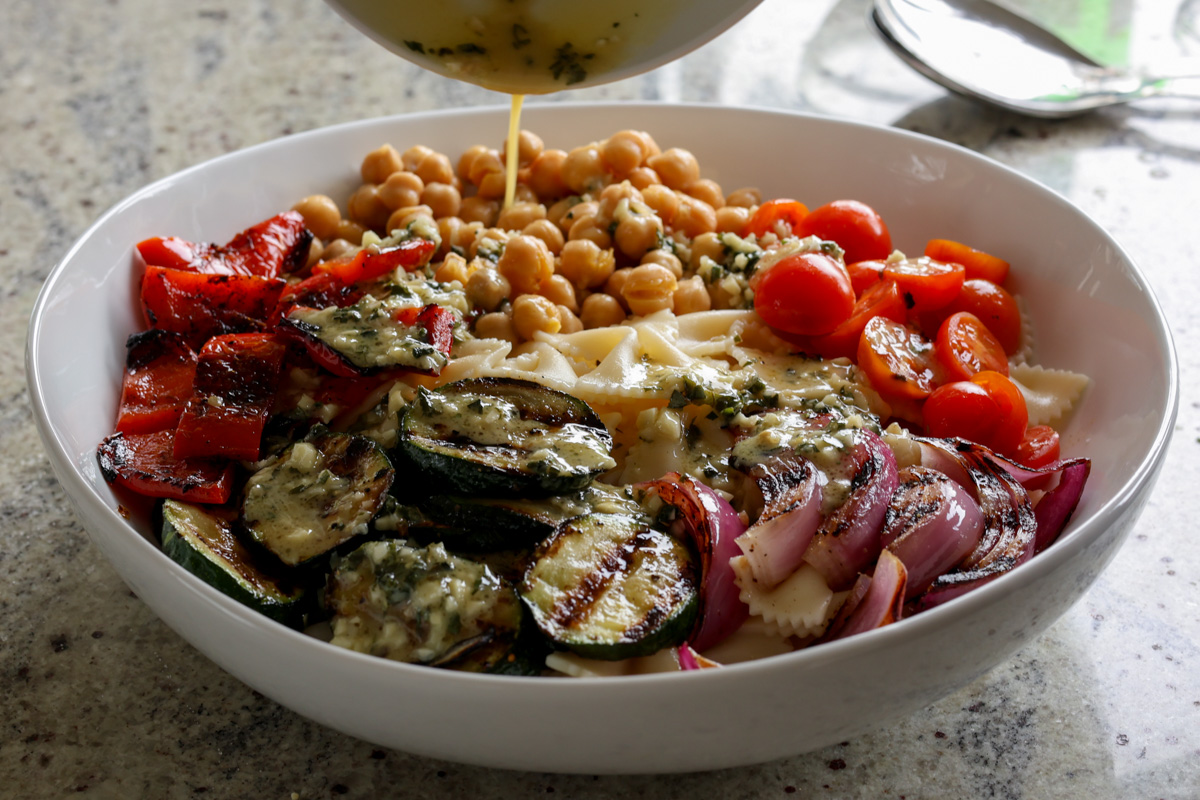 adding Italian dressing to chickpeas, pasta, and grilled vegetables in the mixing bowl before mixing