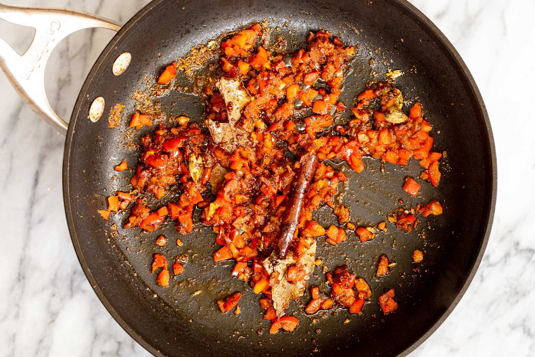 chopped red bell pepper being added to fried spices in a sauteeing pan