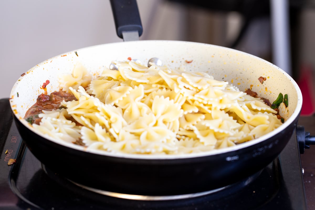 cooked vegan farfalle being added to a pan with sauteed pepperoni pasta sauce