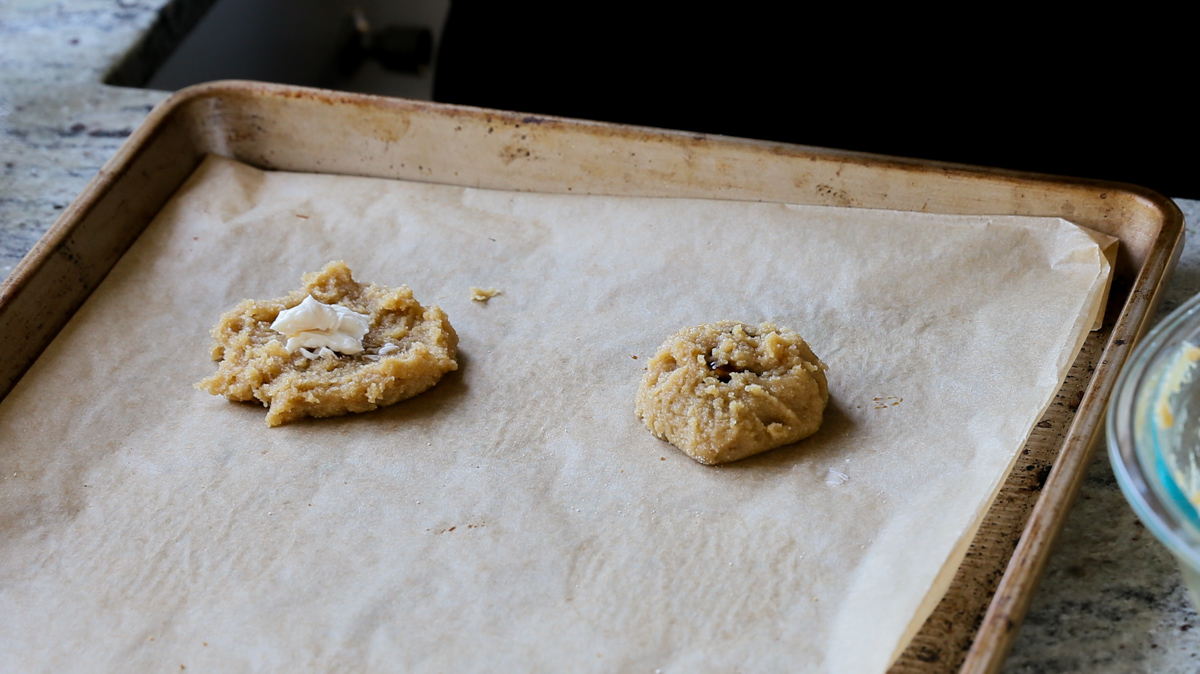 2 unbaked cookies on a baking sheet, one closed up and one with a dollop of cream cheese mixture on it