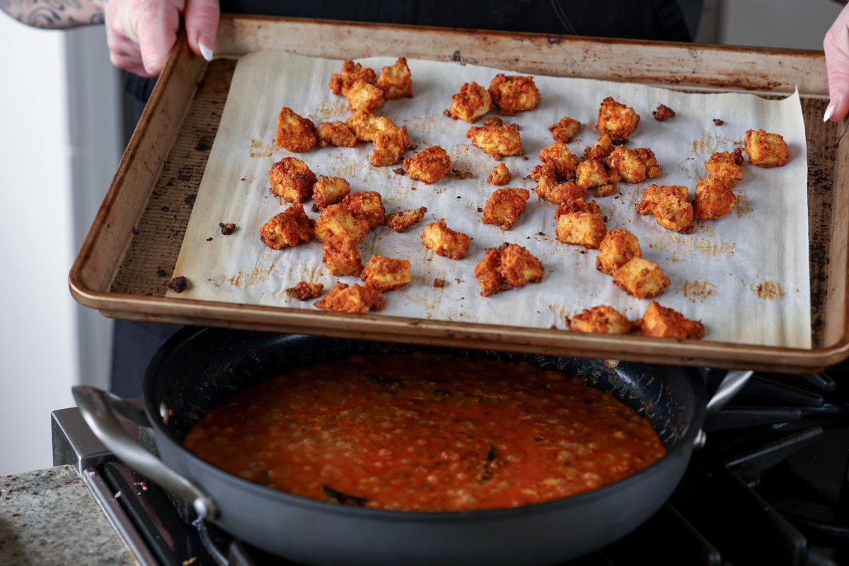 tofu coated in cornstarch and spices on the baking sheet after baking, held over the pan of madurai chicken curry sauce